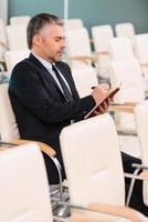 The one and only. Side view of concentrated mature man in formalwear writing something in note pad while sitting on the chair in empty conference hall photo