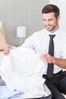 Unpacking his suitcase. Handsome young man in shirt and tie unpacking his suitcase and smiling while sitting on the bed in hotel room photo