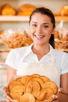 la panadería más fresca para nuestros clientes. Hermosa mujer joven en delantal sosteniendo una cesta con productos horneados mientras está de pie en la panadería. foto