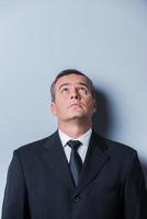 Curious businessman. Curious mature man in formalwear looking up while standing against grey background photo