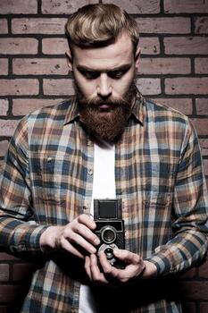 Old-fashioned photographer. Handsome young bearded man holding hands in pockets and looking at camera while standing against grey background photo