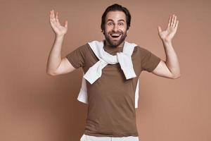 Excited young man gesturing and smiling while standing against brown background photo