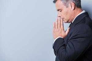 Praying for success. Side view of concentrated mature man in formalwear holding hands clasped near face and keeping eyes closed while standing against grey background photo