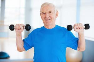 mantenerse saludable. feliz anciano haciendo ejercicio con pesas y sonriendo mientras está de pie en el interior foto