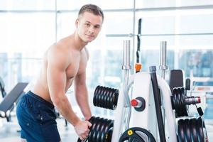 Choosing the right weight. Confident young muscular man choosing weight to lift and smiling while standing in gym photo