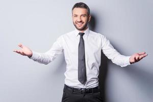 You are welcome Cheerful mature man in shirt and tie gesturing welcome sign and smiling while standing against grey background photo