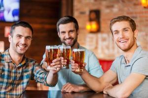 disfrutando de la cerveza con amigos. tres jóvenes felices con ropa informal brindando con cerveza mientras se sientan juntos en el bar foto