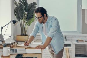 Confident young man making some notes while standing near his working place in office photo