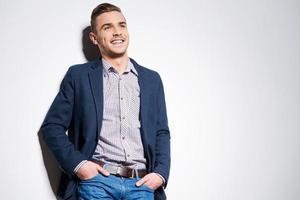 Confident handsome. Handsome young man in smart casual wear holding hands in pockets and smiling while standing against a wall photo