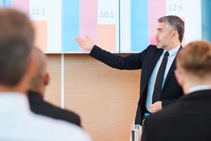 Talking about numbers. Confident mature man in formalwear pointing projection screen with graph on it while making presentation in conference hall with people on the foreground photo