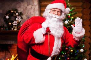 feliz navidad alegre santa claus llevando saco con regalos de navidad y agitando la mano con chimenea y árbol de navidad en el fondo foto