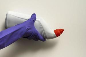 A hand in a purple protective rubber glove holds a plastic white bottle of detergent photo