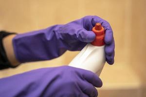 las manos con guantes de goma púrpura sostienen una botella de plástico de detergente y desenroscan la tapa roja foto