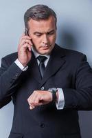 He knows that time is money. Confident mature man in formalwear checking the time and talking on the mobile phone while standing against grey background photo