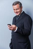 Good business news. Confident mature man in formalwear holding mobile phone and looking at it with smile while standing against grey background photo