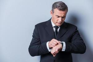 Businessman checking the time. Confident mature man in formalwear checking the time while looking at his watch and standing against grey background photo