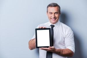 copie el espacio en su tableta. hombre maduro confiado en camisa y corbata mostrando su tableta digital y sonriendo mientras está de pie contra el fondo gris foto