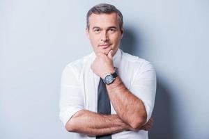 Thinking about your problems. Thoughtful mature man in shirt and tie holding hand on chin and looking at camera while standing against grey background photo