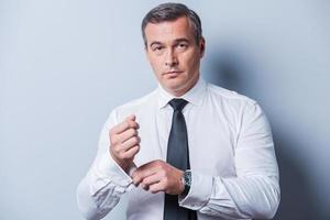 Making business look good. Confident mature man in shirt and tie adjusting his sleeves and looking at camera  while standing against grey background photo