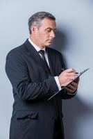 Businessman making notes. Side view of confident mature man in formalwear writing something in clipboard and looking at camera while standing against grey background photo