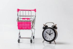 Small supermarket grocery push cart for shopping toy with wheels and classic alarm clock isolated on white background. Sale time buy mall market shop consumer concept. Copy space. photo