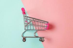 Small supermarket grocery push cart for shopping toy with wheels isolated on blue and pink pastel colorful paper trendy geometric background Copy space. Sale buy mall market shop consumer concept. photo