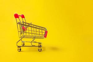 Small supermarket grocery push cart for shopping toy with wheels isolated on yellow colourful trendy modern fashion background. Sale buy mall market shop consumer concept. Copy space. photo