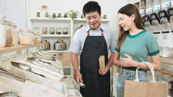 serviceperson im einzelhandelsgeschäft, asiatischer männlicher ladenbesitzer, der im nachfüllladen mit einer einkaufstüte aus recyceltem papier, abfallfreie lebensmittel, bio-produkte, nachhaltiger lebensstil snacks an kunden schöpft. video