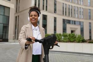 a young American woman marketer with a wide smile stands on an electric scooter in headphones near a business center photo