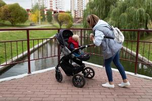 una joven madre con una mochila en los hombros camina con su hijo en un cochecito en el parque de la ciudad foto