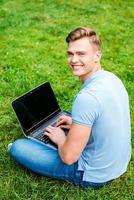 Surfing the net wherever I want. Top view of happy young man looking over shoulder and smiling while sitting on the grass with laptop photo