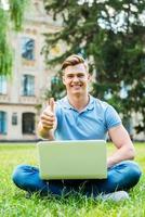 estudiante exitoso un joven feliz mirando la cámara y mostrando su pulgar hacia arriba mientras se sienta en el césped con una laptop foto