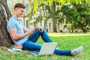 relajarse después de clase. vista lateral de un apuesto estudiante universitario masculino escuchando un reproductor de mp3 mientras se sienta en el césped y se apoya en el árbol foto