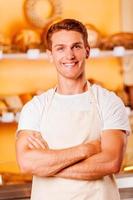 Confident baker. Handsome young man in apron keeping arms crossed and smiling while standing in bakery shop photo