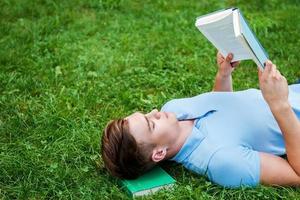 Finding a peaceful place to read. Top view of handsome young man reading a book while lying on grass with laptop photo