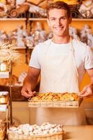 The freshest cookies for you. Handsome young man in apron holding tray with cookies and smiling while standing in bakery shop photo