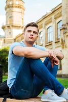 pensando en el futuro. estudiante masculino pensativo sentado en el banco y mirando hacia otro lado con el edificio de la universidad en el fondo foto