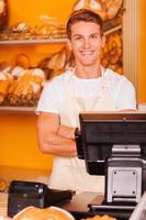 cajero en panadería. apuesto joven cajero masculino en delantal con los brazos cruzados y sonriendo mientras está de pie en la caja registradora en la panadería foto