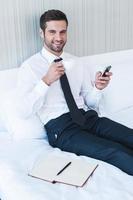 Typing business message. Cheerful young man in shirt and tie drinking coffee and holding mobile phone while lying in bed at the hotel room photo