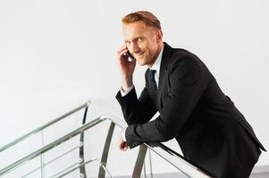 Business on the phone. Side view of confident young man in formalwear leaning at the metal handrail and talking on the mobile phone photo