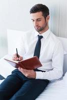 Making some urgent notes. Confident young man in shirt and tie writing something in note pad while sitting in bed at the hotel room photo