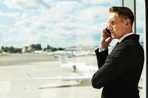 Businessman waiting for flight. Rear view of thoughtful businessman in formalwear looking through a window and talking on the mobile phone while standing in airport photo