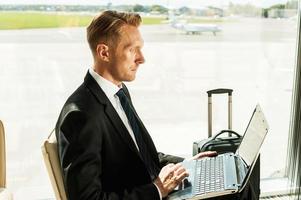 Taking advantages of free Wi-Fi. Side view of confident businessman in formalwear working on laptop while waiting for a flight in airport photo