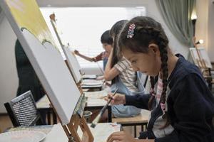 A little girl concentrates on acrylic color picture painting on canvas with multiracial kids in an art classroom, creative learning with talents and skills in the elementary school studio education. photo