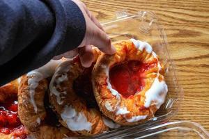 A man's hand is holding a Cherry filled Danish or Danish bread in a plastic box and placed it on a brown wooden table. photo