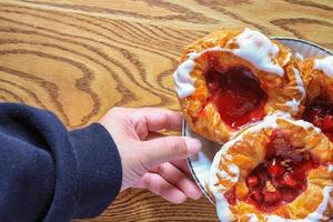 Man's hand holding Cherry filled Danish or Danish bread served in a white plate on a brown wooden table. photo