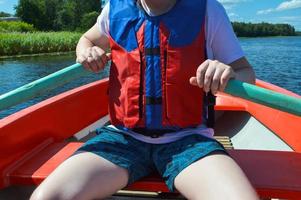 un hombre en un bote rojo, pantalones cortos y un chaleco salvavidas está remando con remos en un bote para dar un paseo por el agua del lago río mar en la naturaleza foto