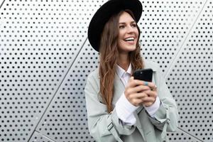 retrato de una chica fuerte con un abrigo de otoño y un sombrero negro contra el fondo de una pared de aluminio de un edificio con un teléfono móvil en las manos, concepto de trabajo independiente y remoto foto