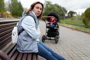 madre joven molesta sentada en el parque en un banco lejos del cochecito de bebé con una cara enojada. el concepto de depresión posparto foto