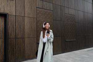 portrait of a stylish young woman in headphones using a mobile phone against the background of a brown wall of a modern building photo
